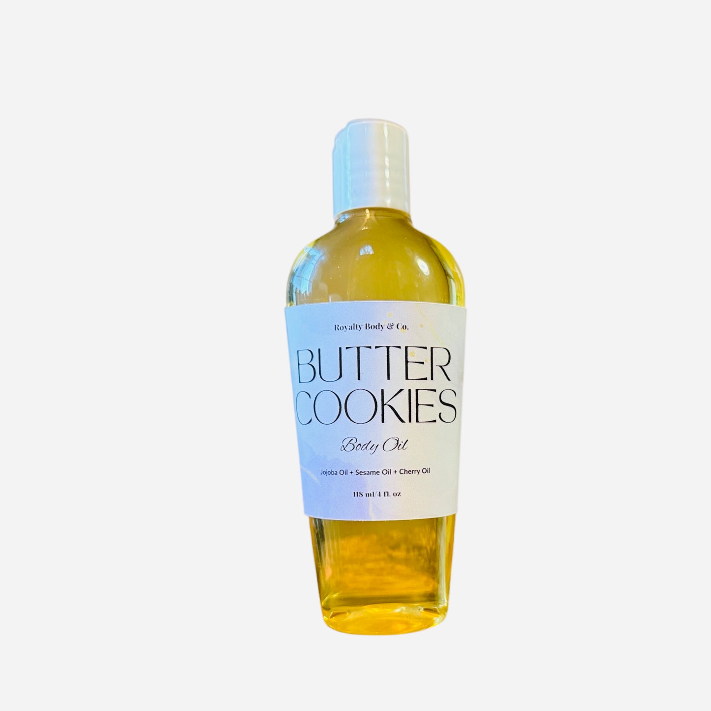 The clear bottle of Butter Cookies Body Oil contains yellow-tinted oil promising intense hydration with jojoba, sesame, and cherry oils. It features a minimalistic design with a white cap and ensures long-lasting fragrance, all set against a plain white background.