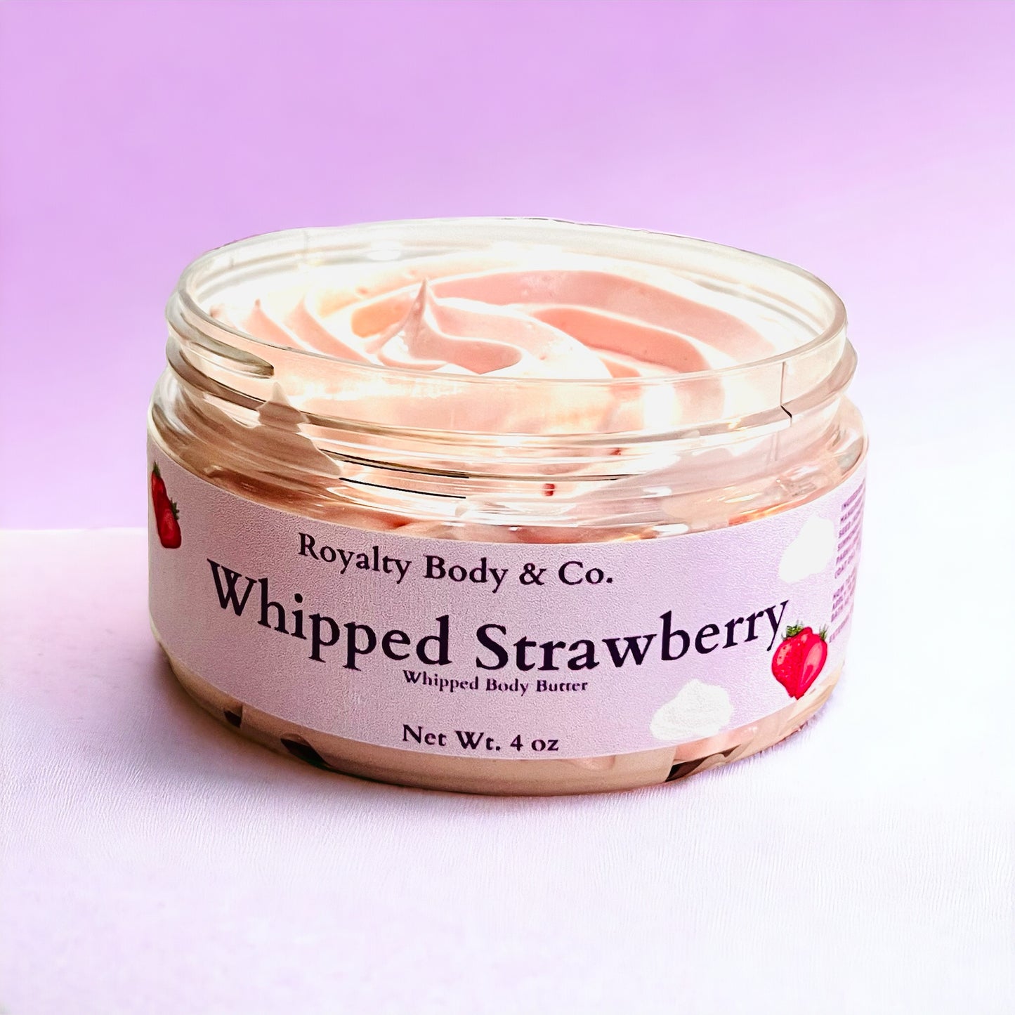 An open jar of 4 oz Whipped Strawberry Body Butter from Royalty Body & Co. sits against a gradient pink backdrop. The label features strawberries and clouds, ideal for eczema-prone skin needing deep hydration. Inside is a luxurious swirl of whipped butter.