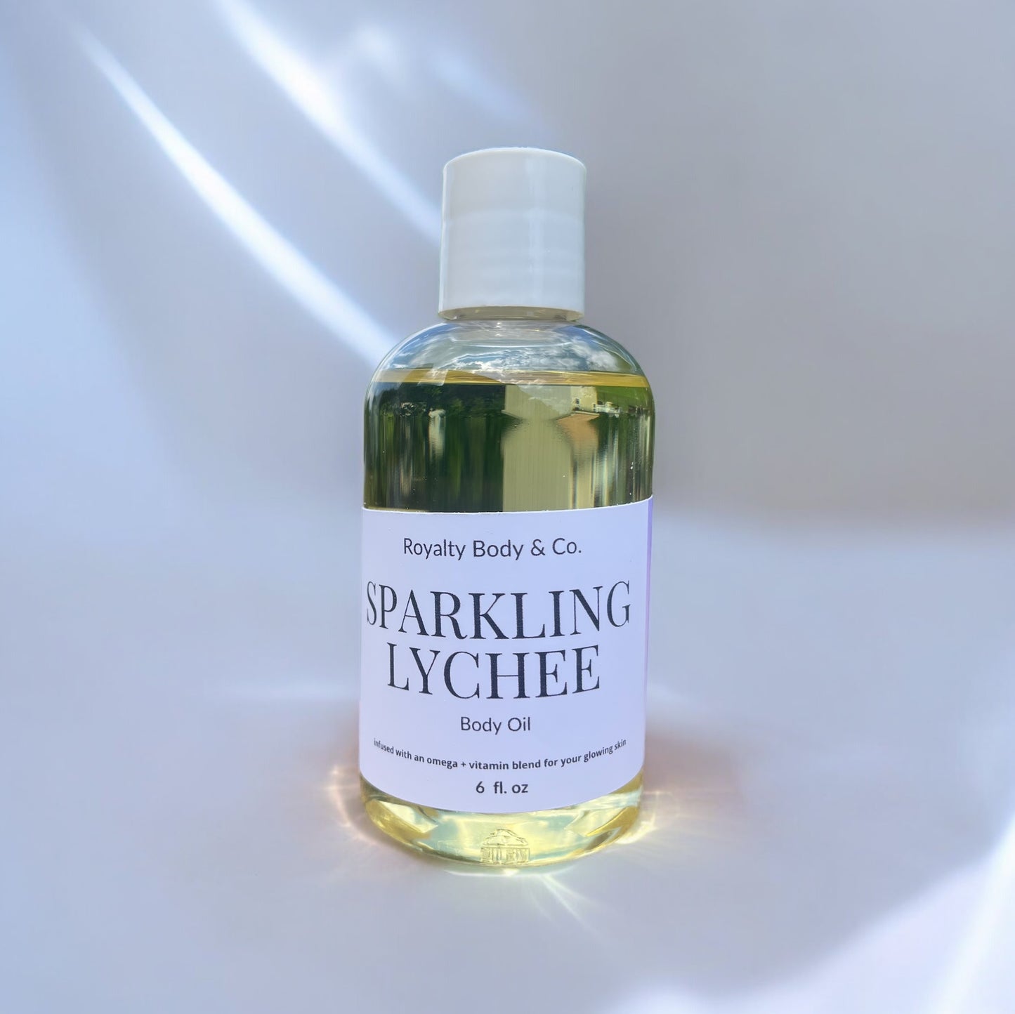 The Sparkling Lychee Body Oil by Royalty Body & Co. features a gentle formula ideal for eczema-prone skin, packaged in a bottle with a white cap and containing nourishing pale yellow liquid presented against a softly lit background.