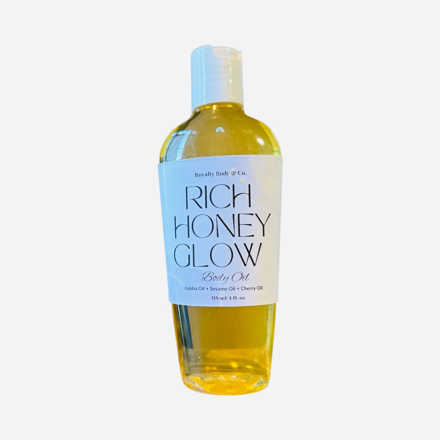 The clear bottle of Rich Honey Glow Body Oil features a white label, revealing a golden-yellow liquid for deep hydration. It contains Jojoba, Sesame, and Cherry Oils. The packaging evokes honey butter and is set against a simple white backdrop.