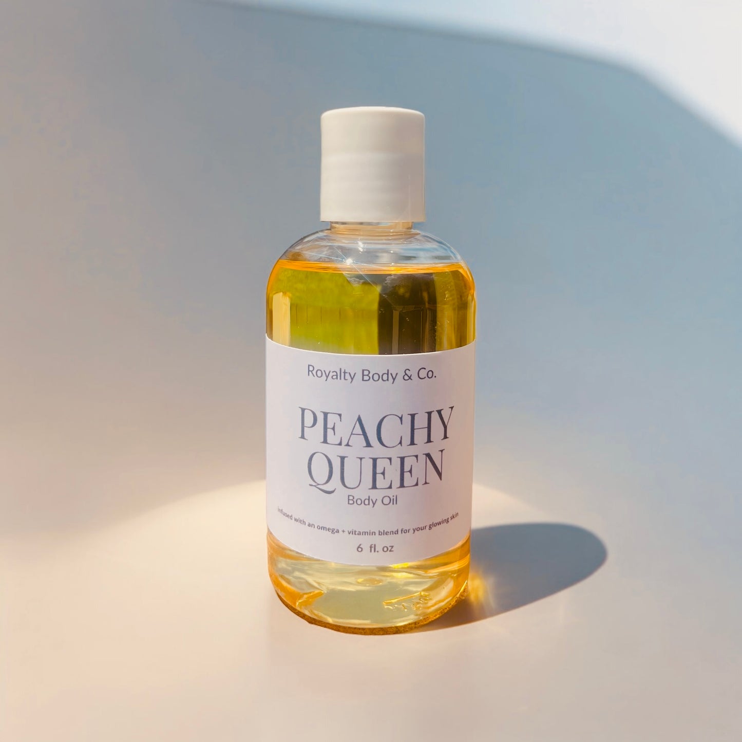 A clear bottle of hydrating oil in a yellow hue, capped with white, displays the label Royalty Body & Co. PEACHY QUEEN Body Oil. It features an exotic scent and omega-vitamin blend details on a reflective surface against a light gradient backdrop.