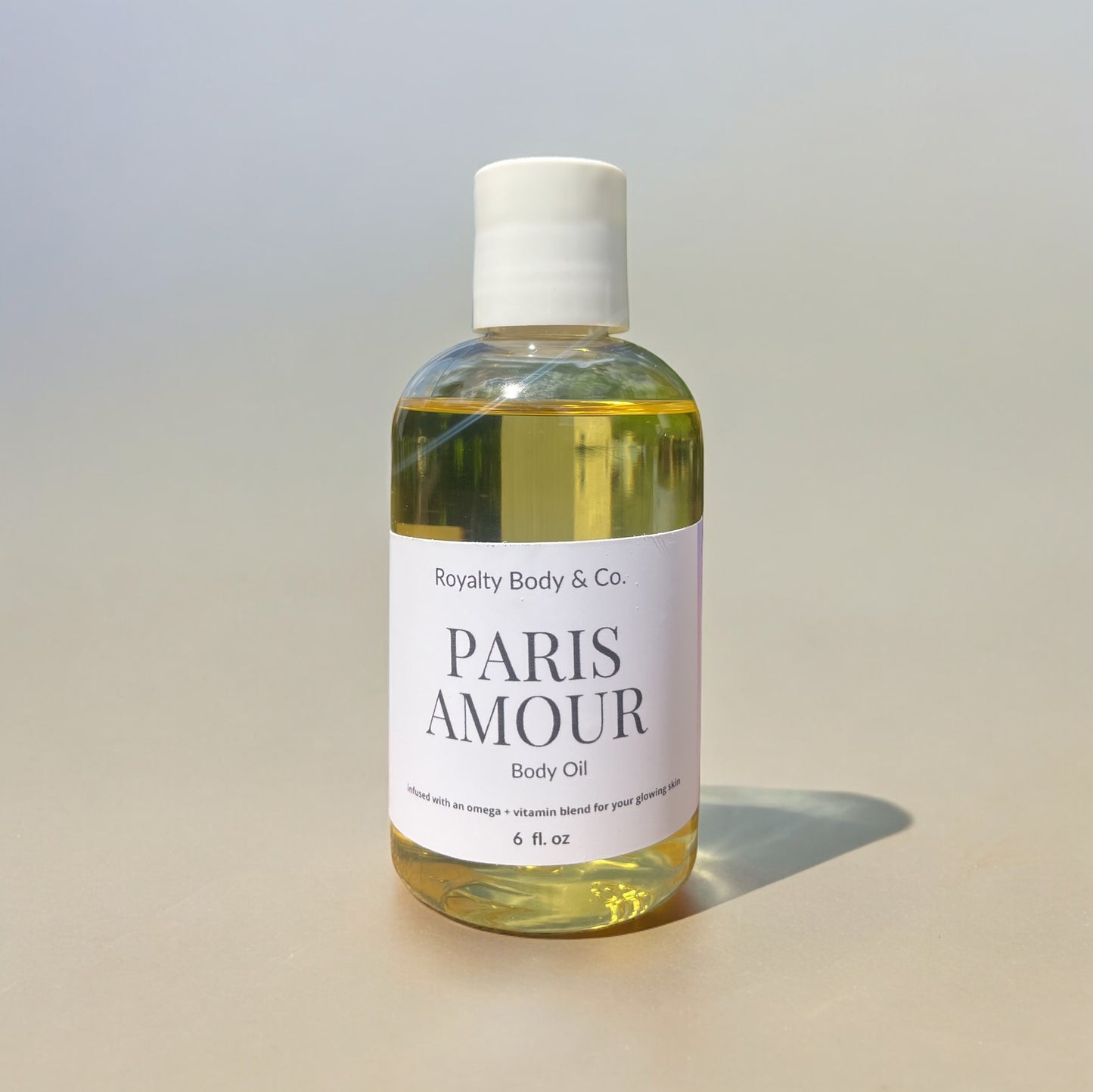 A clear 6-fl-oz bottle of Paris Amour Body Oil from Royalty Body & Co. sits on a soft, neutral background, featuring a nourishing blend of omegas and vitamins with an alluring fragrance to enhance your skincare ritual.