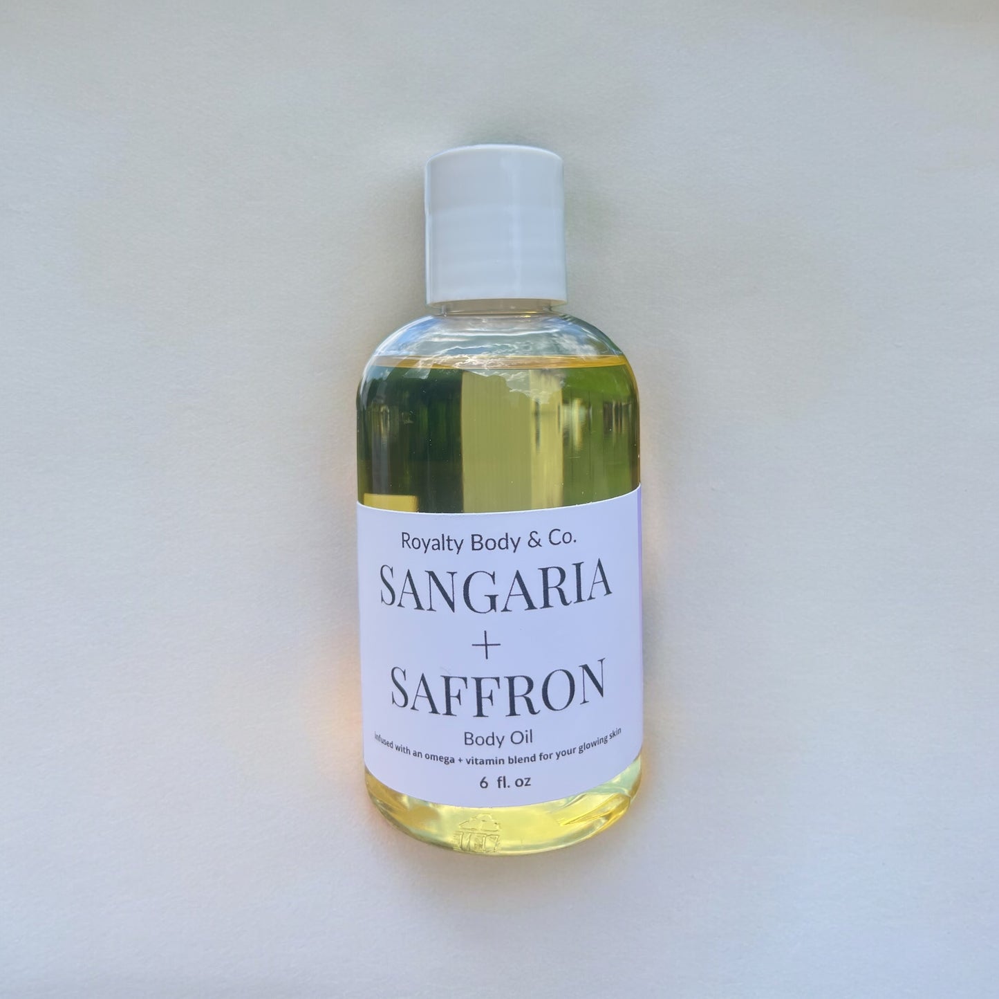 A clear plastic bottle labeled Sangria + Saffron Body Oil from Royalty Body & Co. holds 6 fl. oz of yellow nourishing oil. Its an omega and vitamin blend designed for glowing, hydrated skin and is showcased on a light, plain background.