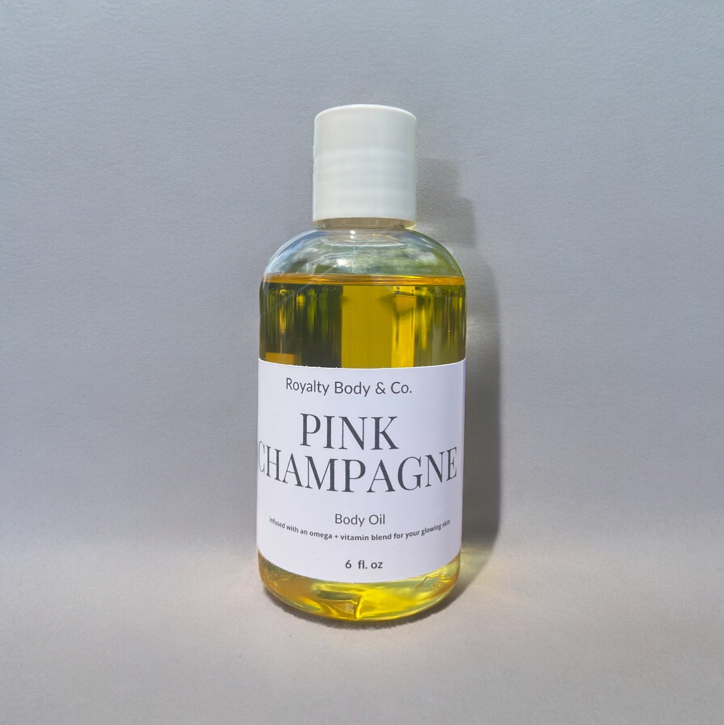 The Pink Champagne Body Oil by Royalty Body & Co. is a clear bottle with a white cap. It holds 6 fl oz of light yellow oil, featuring an omega and vitamin blend ideal for sensitive skin, perfect for nourishing dry skin against a neutral background.